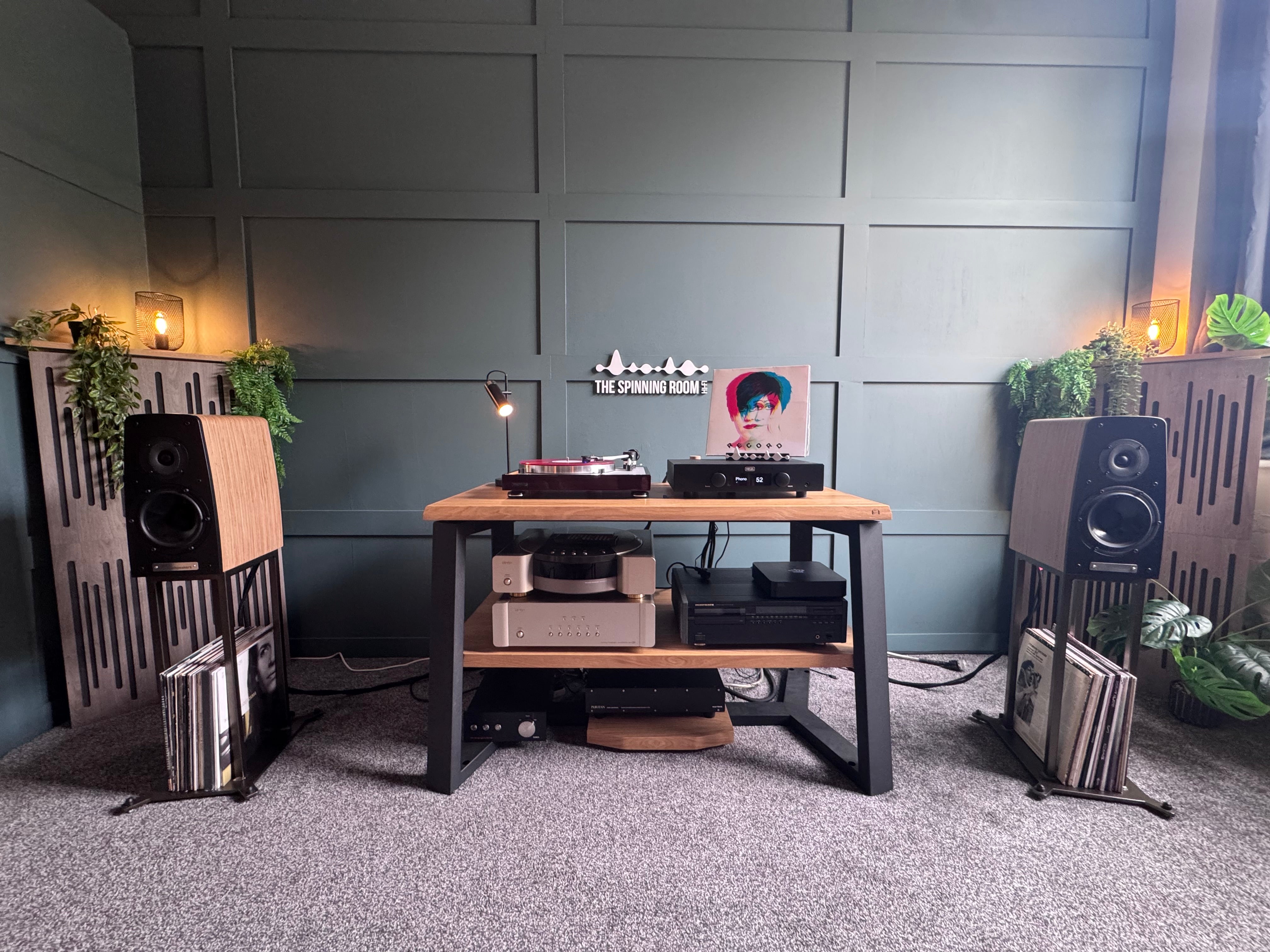 A styled home audio setup in a room with paneled gray walls and a gray carpet, featuring a wooden console table with audio equipment, two large speakers on stands, and vinyl records.
